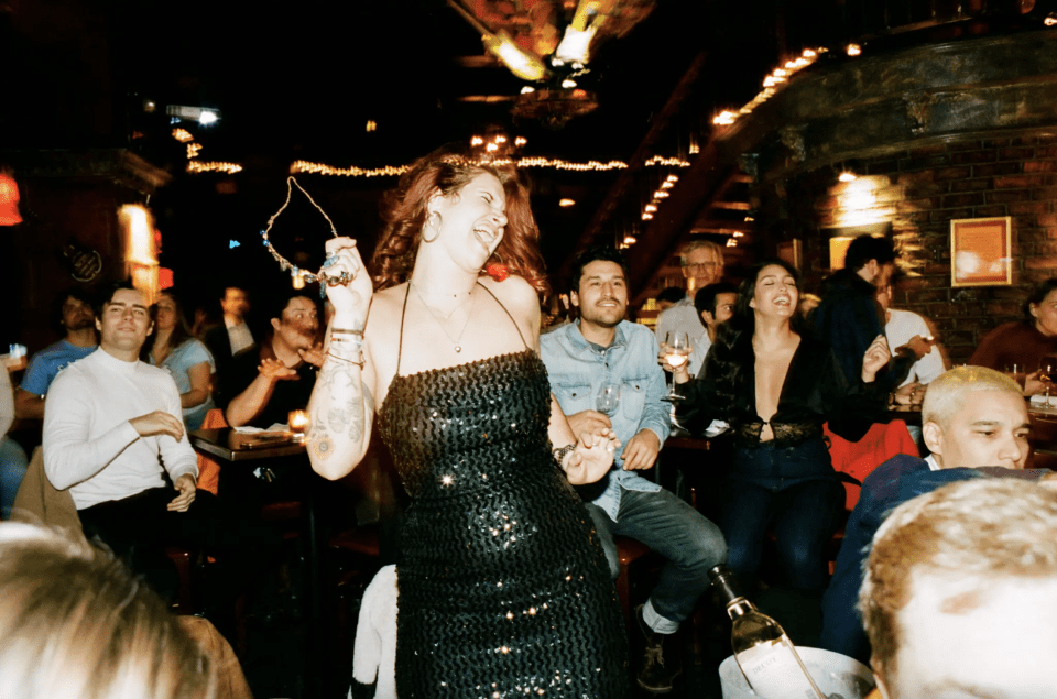 A woman in a black dress dances in front of friends at a dueling pianos show.