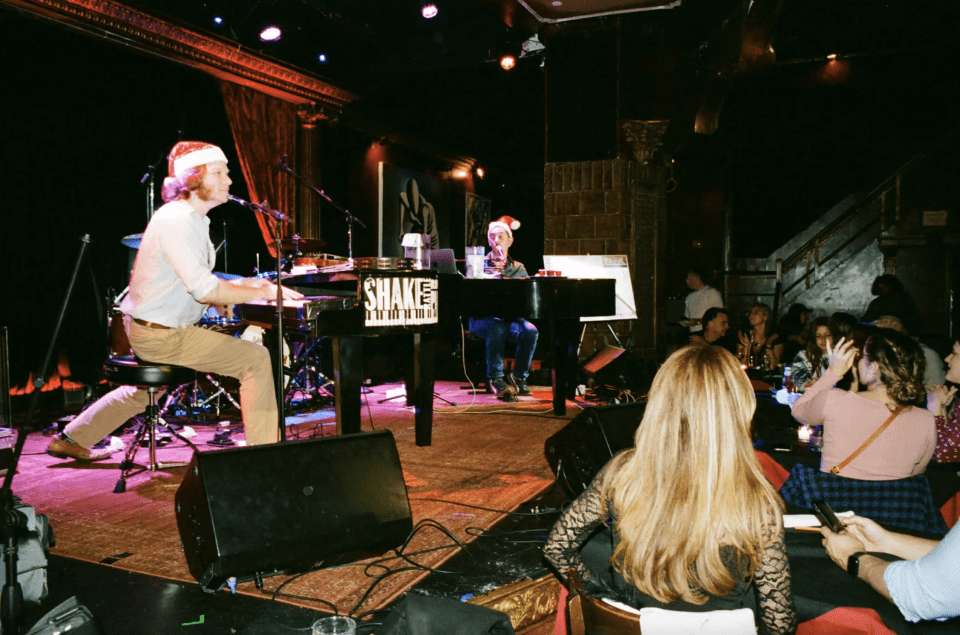 Two pianists in Santa hats sing to the crowd at a dueling pianos show.