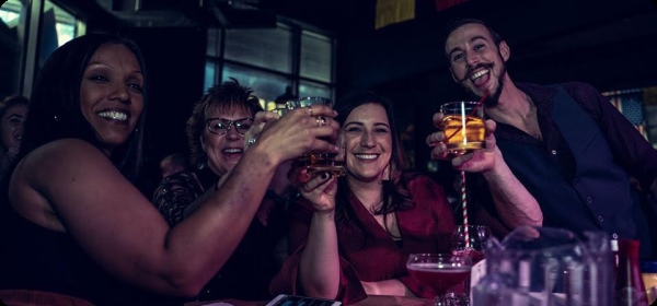 Three smiling women and a piano player toast the camera at a Boozy Brunch show.