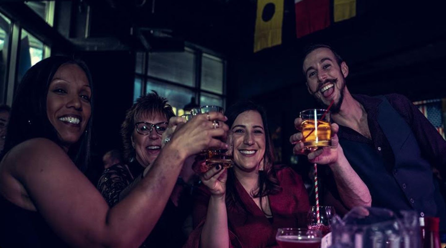 Three smiling women and a piano player toast the camera at a Boozy Brunch show.