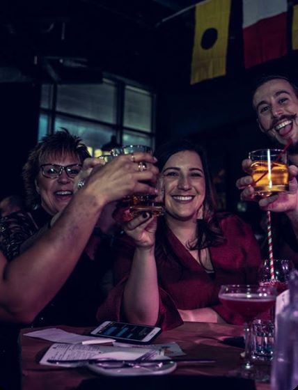 Three smiling women and a piano player toast the camera at a Boozy Brunch show.