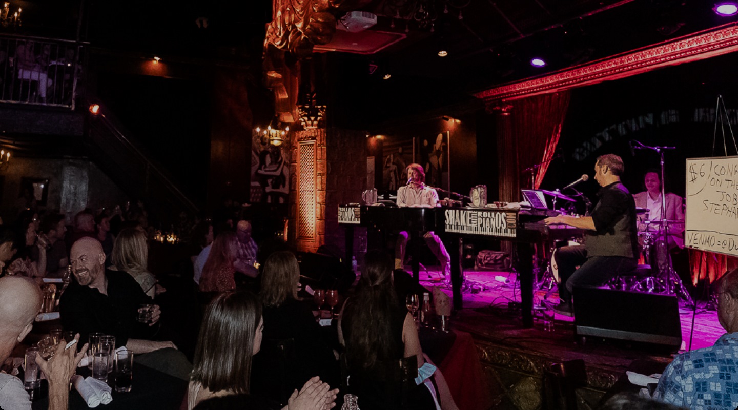 2 Dueling pianists play on a multicolored stage to a packed crowd at a Saturday night show in NYC.