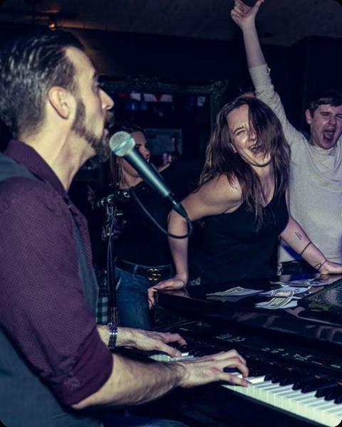 A pianist sings while a young woman and two men do a lively dance alongside the piano.