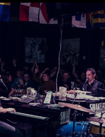 Mark singing at the piano while a crowd cheers. There are a number of colorful flags in the bar.