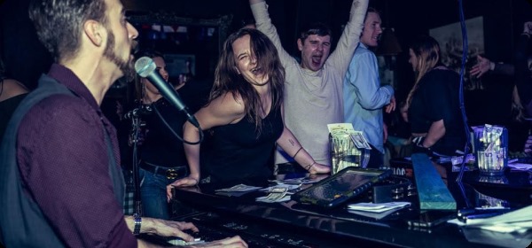 A pianist sings while a young woman and two men do a lively dance alongside the piano.